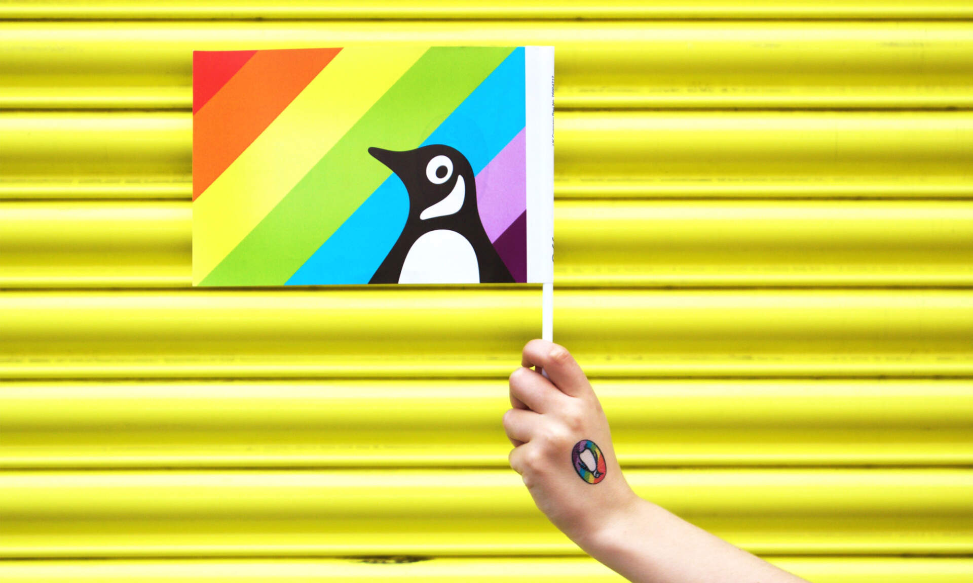 Penguin Pride flag held by a hand wearing a branded temporary tattoo.